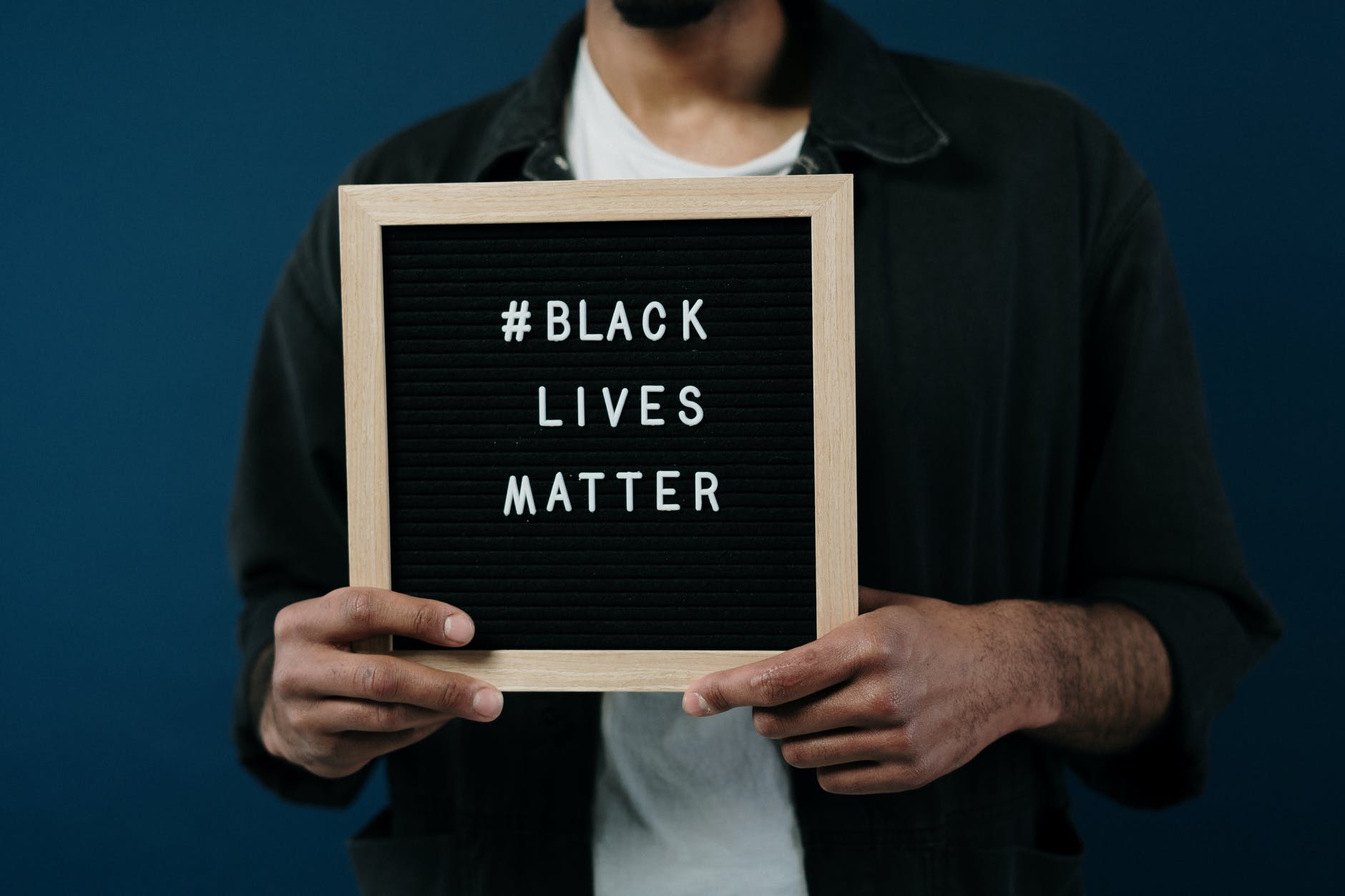 Photo of a Black man's torso holding a board that says # Black Lives Matter in his hands